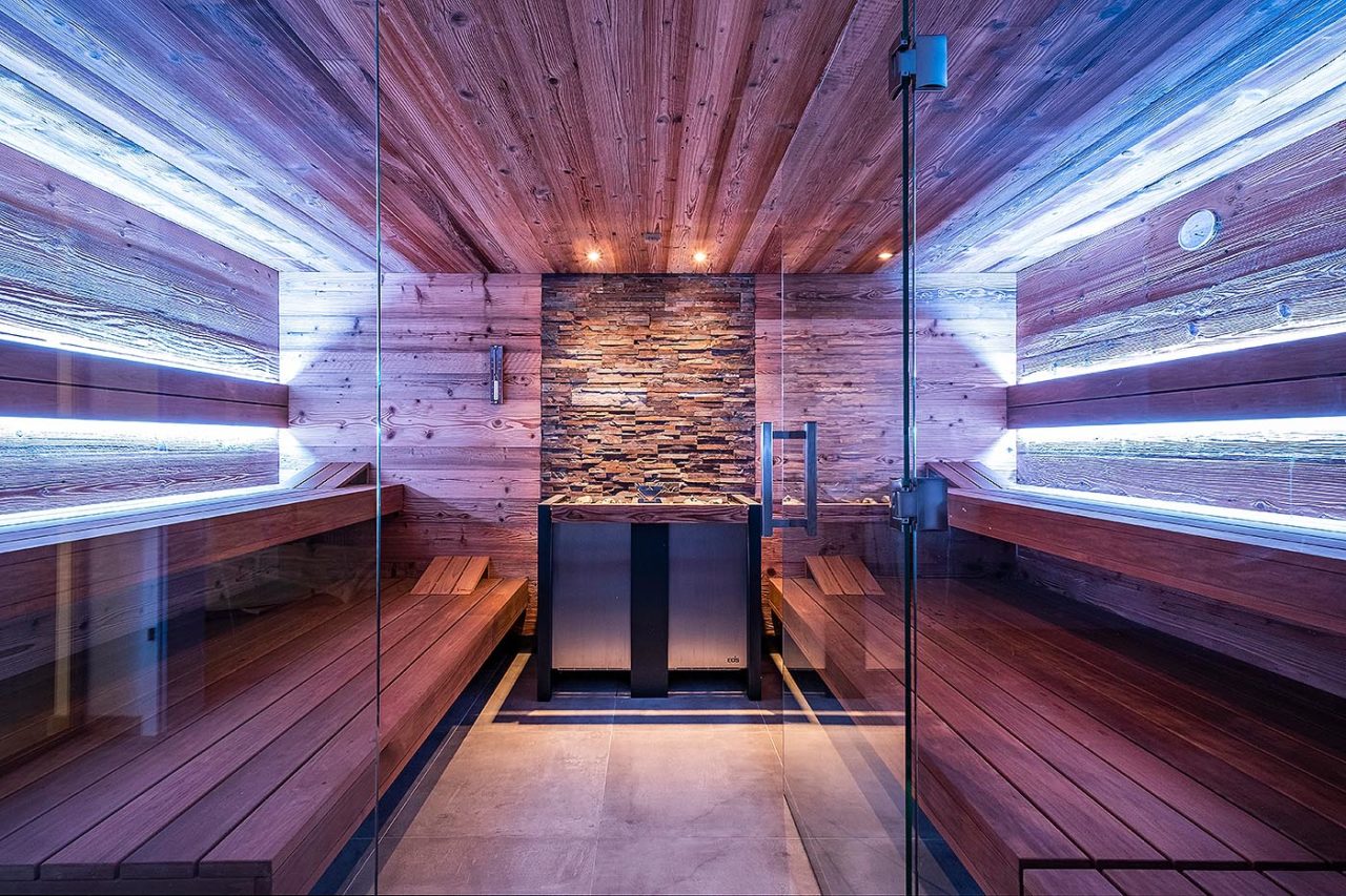 Luxus Sauna: Design Sauna Zuhause: Moderne Sauna aus Altholz mit symmetrischen Linien, Steinwand hinter dem großen Saunaofen und indirekter LED Beleuchtung.