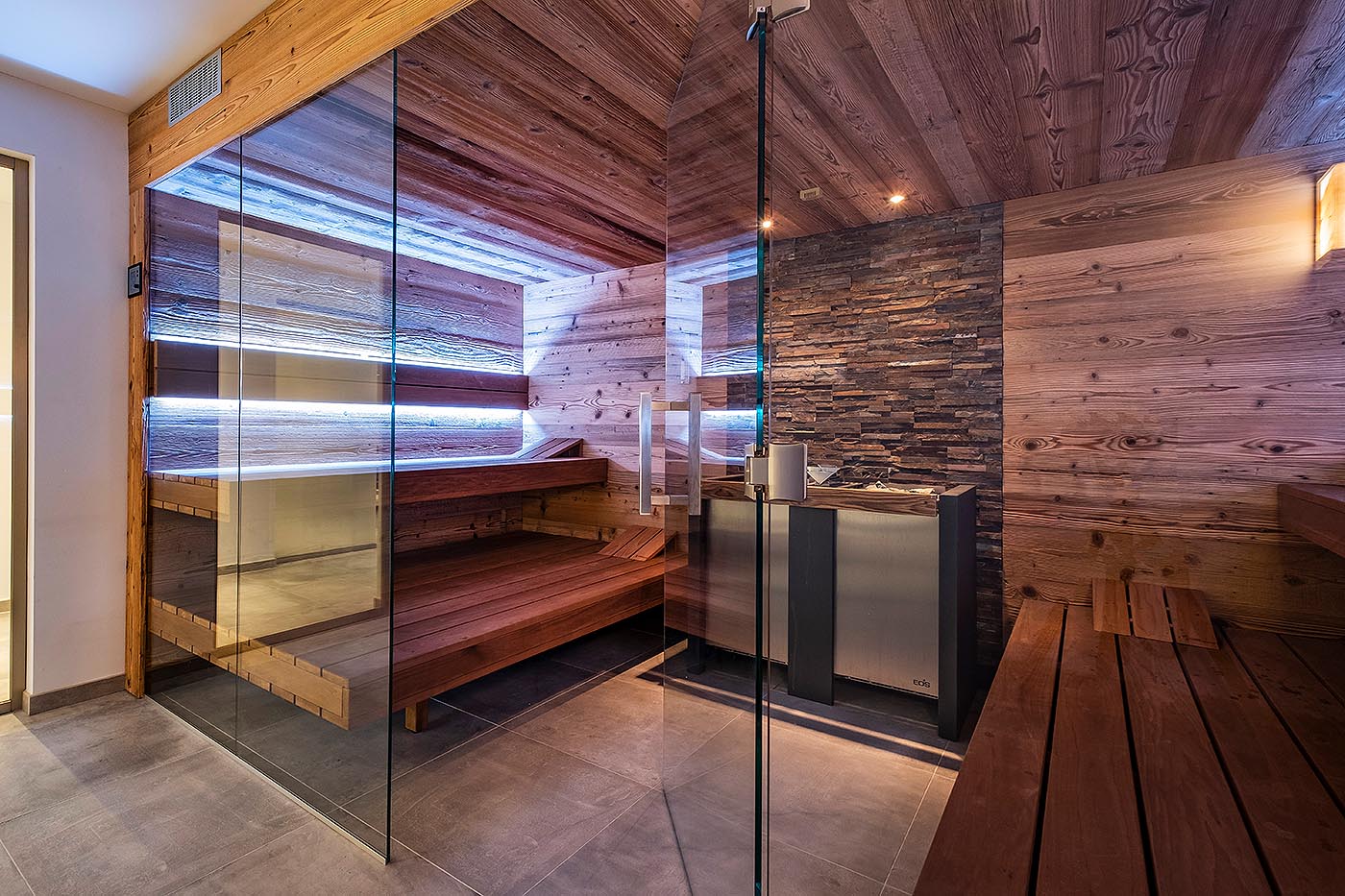 Altholz Design Sauna für Zuhause mit Glasfront und dekorativer Steinwand hinter dem großen Saunaofen. Schwebende Saunabänke, indirekte Beleuchtung.