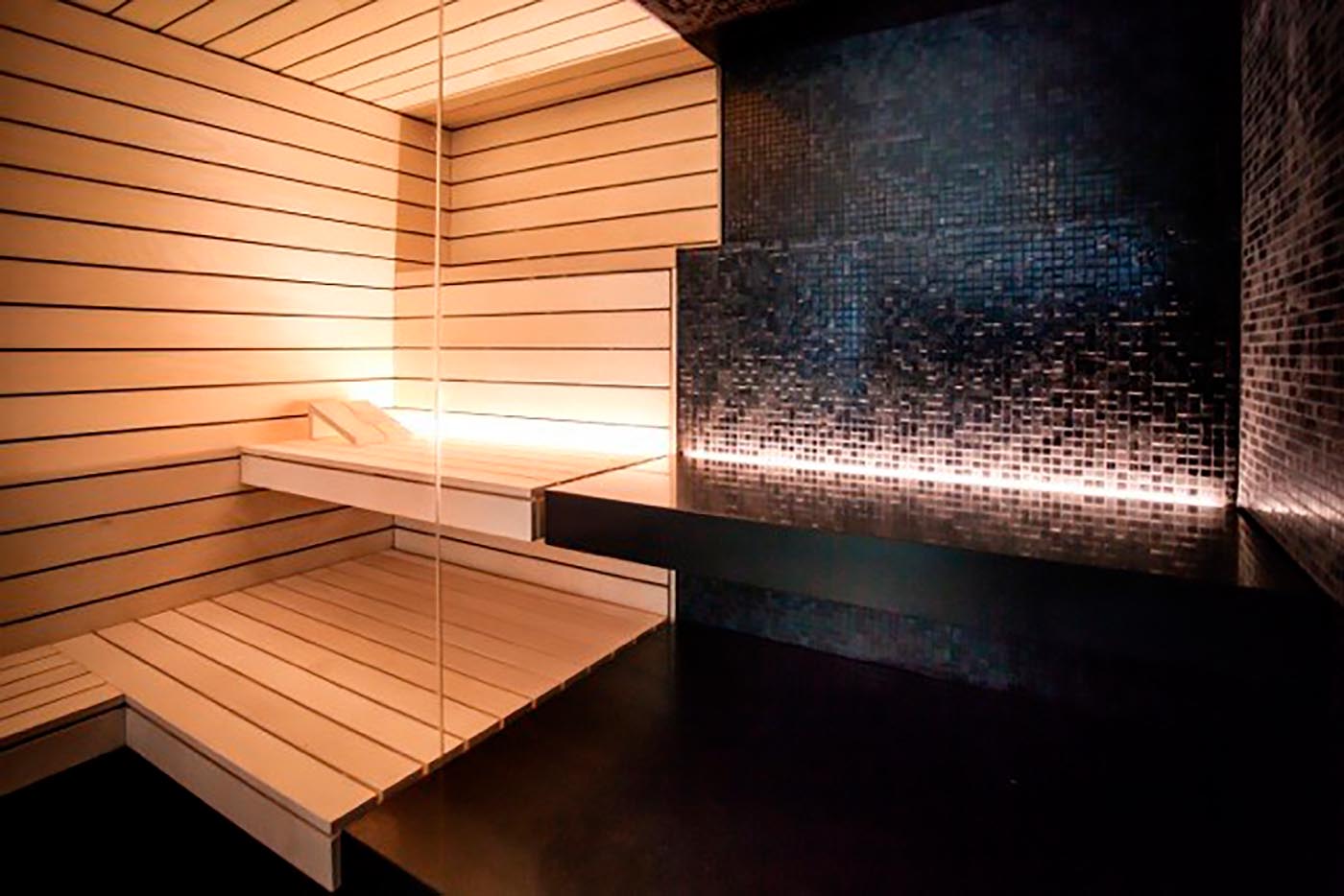 Kombination aus einer hellen Sauna mit Hinterwandofen und direkt daneben einem Dampfbad in schwarzen Mosaikfliesen, nur getrennt durch eine Glasscheibe