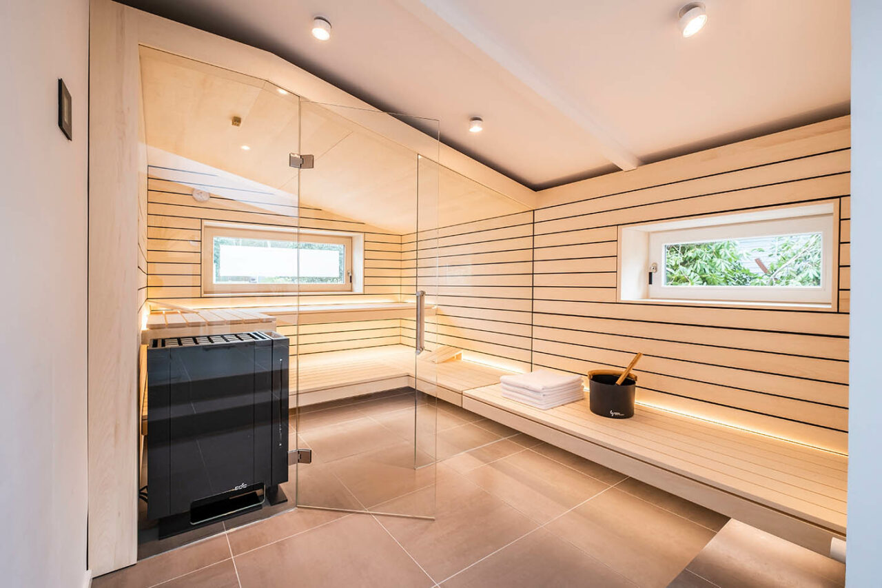 Design Sauna mit Fenster nach draußen. Sauna mit Dachschräge nach Maß in hellem Holz. Große Glasfront, nach außen verlängerte Bänke und LED Beleuchtung. Frei stehender Saunaofen und schwebende Saunabänke.