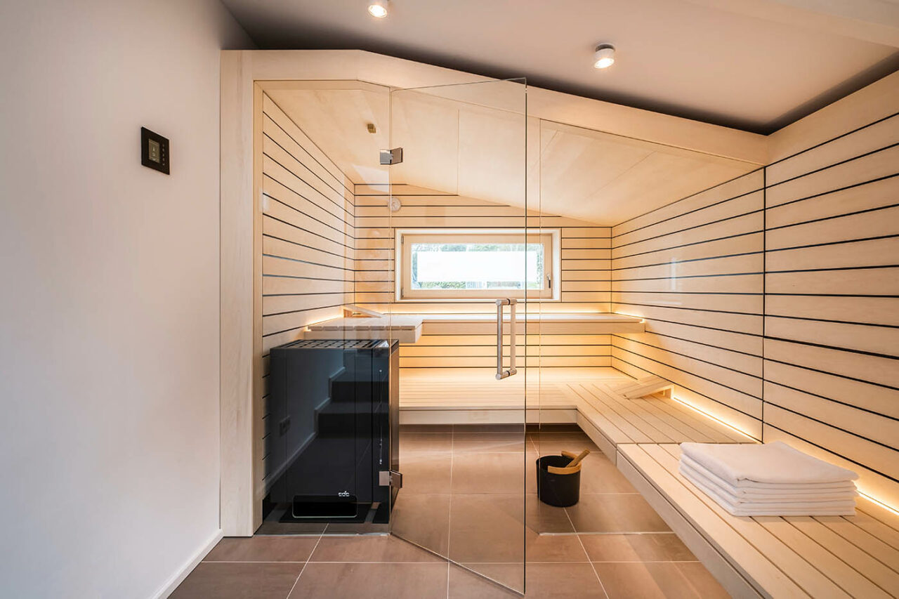 Design Sauna mit Fenster nach draußen. Sauna mit Dachschräge nach Maß in hellem Holz. Große Glasfront, nach außen verlängerte Bänke und LED Beleuchtung. Frei stehender Saunaofen und schwebende Saunabänke.