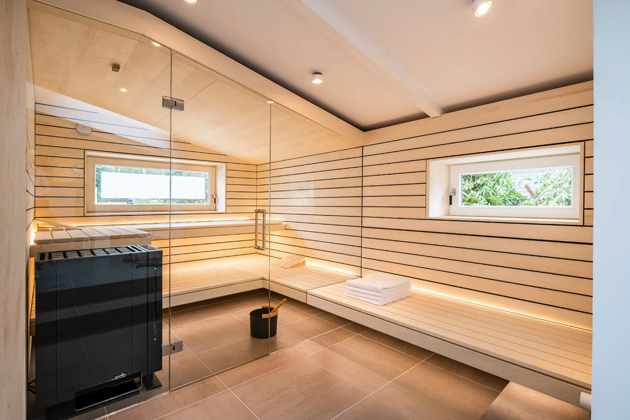 Design Sauna mit Fenster nach draußen. Sauna mit Dachschräge nach Maß in hellem Holz. Große Glasfront, nach außen verlängerte Bänke und LED Beleuchtung. Frei stehender Saunaofen und schwebende Saunabänke.