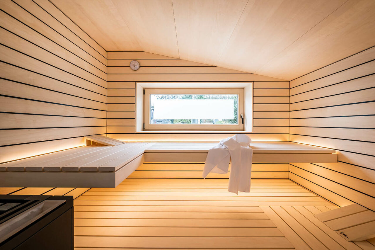 Design Sauna mit Fenster nach draußen. Sauna mit Dachschräge nach Maß in hellem Holz. Große Glasfront, nach außen verlängerte Bänke und LED Beleuchtung. Frei stehender Saunaofen und schwebende Saunabänke.