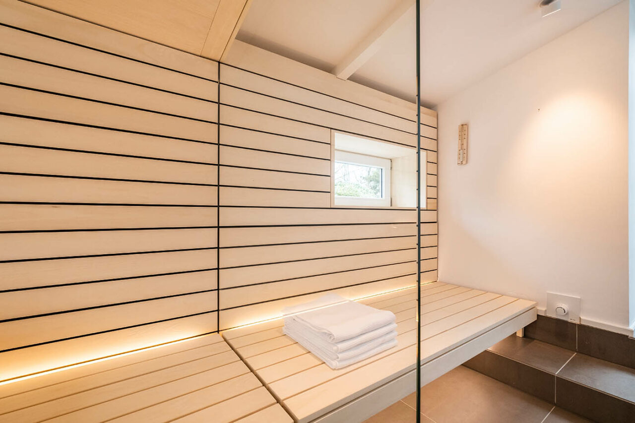 Design Sauna mit Fenster nach draußen. Sauna mit Dachschräge nach Maß in hellem Holz. Große Glasfront, nach außen verlängerte Bänke und LED Beleuchtung. Frei stehender Saunaofen und schwebende Saunabänke.