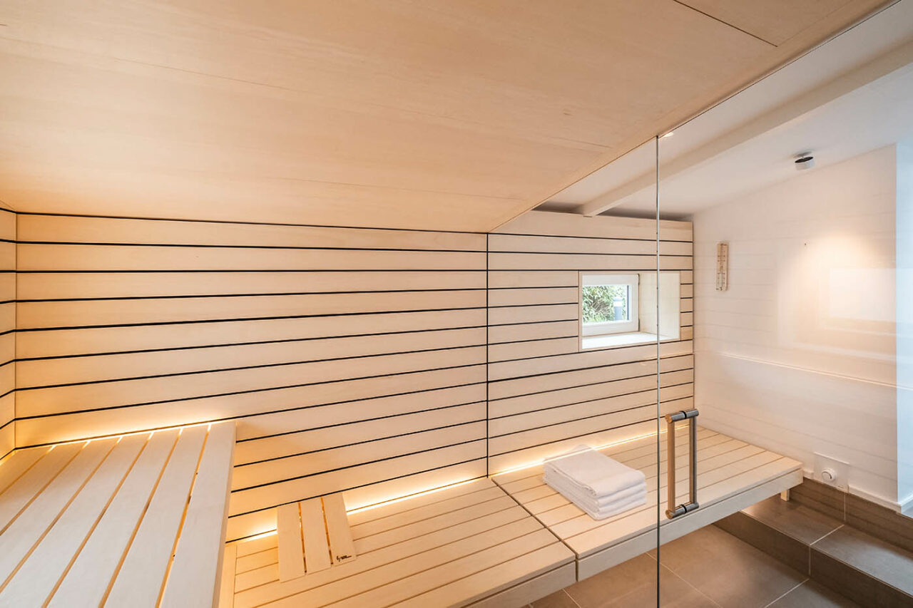 Design Sauna mit Fenster nach draußen. Sauna mit Dachschräge nach Maß in hellem Holz. Große Glasfront, nach außen verlängerte Bänke und LED Beleuchtung. Frei stehender Saunaofen und schwebende Saunabänke.