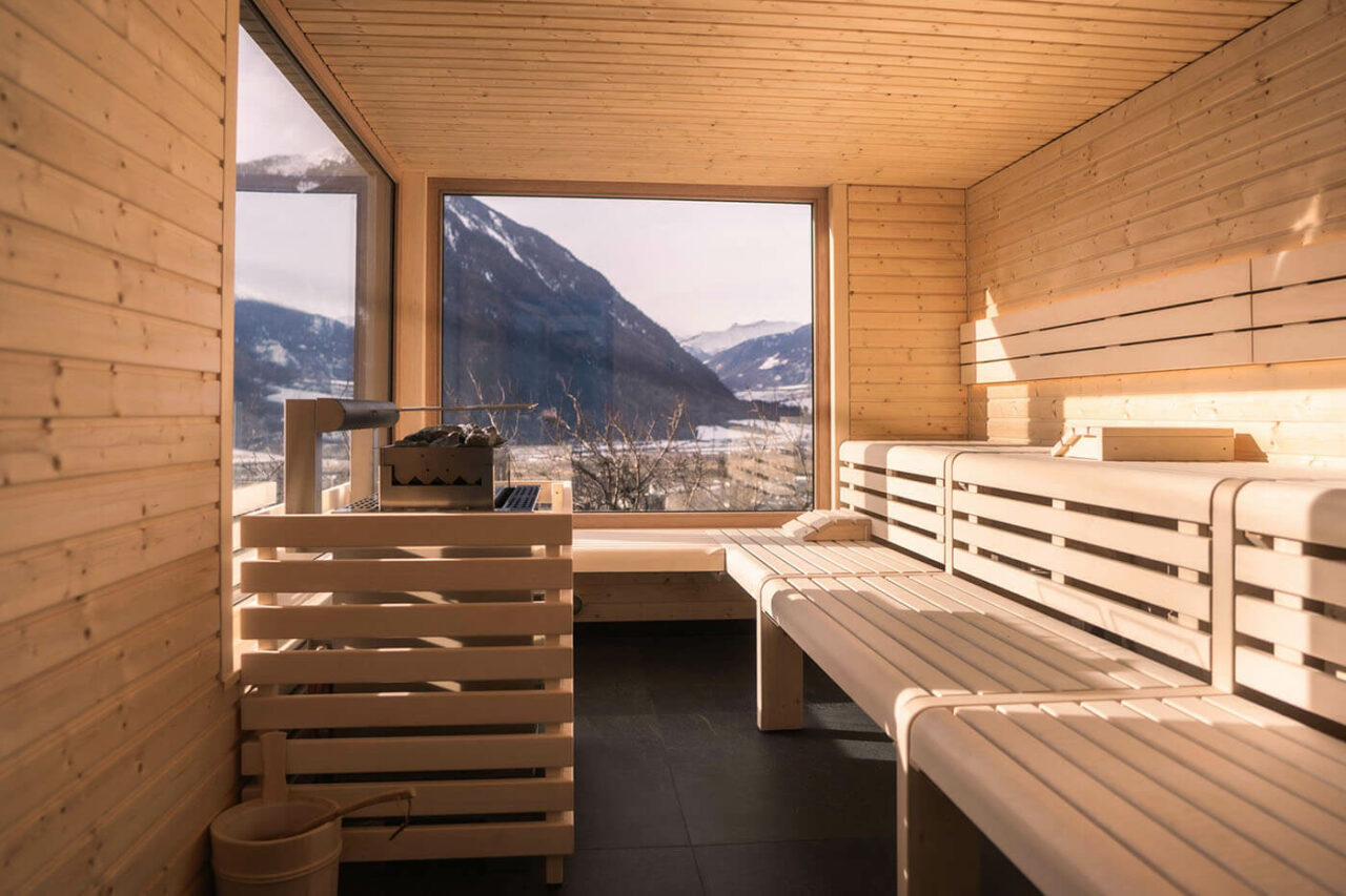 Panoramasauna: Außensauna in Südtirol, Biohotel Panorama. Außenverkleidung in Lärchenholz, helle Inneneinrichtung in Fichte und Espe. Große Glasflächen mit Blick auf die Südtiroler Bergwelt. Saunaofen mit Sicherheitseinrichtung.