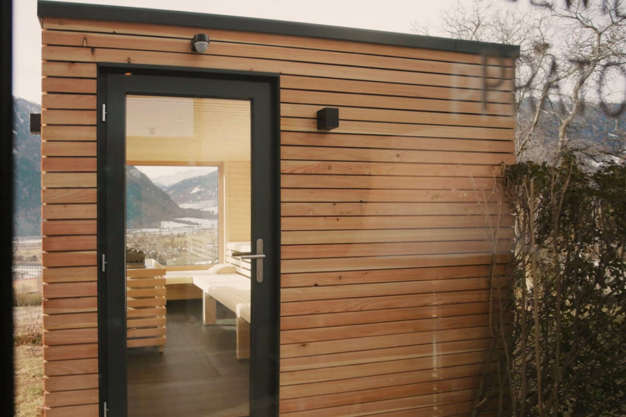 Panoramasauna: Außensauna in Südtirol, Biohotel Panorama. Außenverkleidung in Lärchenholz, helle Inneneinrichtung, verglaste Tür und große Glasflächen mit Blick auf die Südtiroler Bergwelt.