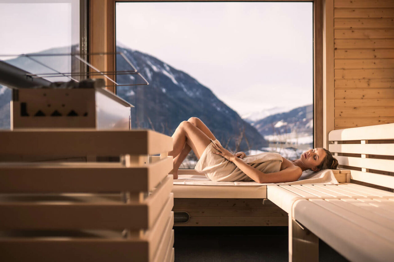 Panoramasauna: Außensauna in Südtirol, Biohotel Panorama. Außenverkleidung in Lärchenholz, helle Inneneinrichtung, verglaste Tür und große Glasflächen mit Blick auf die Südtiroler Bergwelt. Model liegt auf der Saunabank.